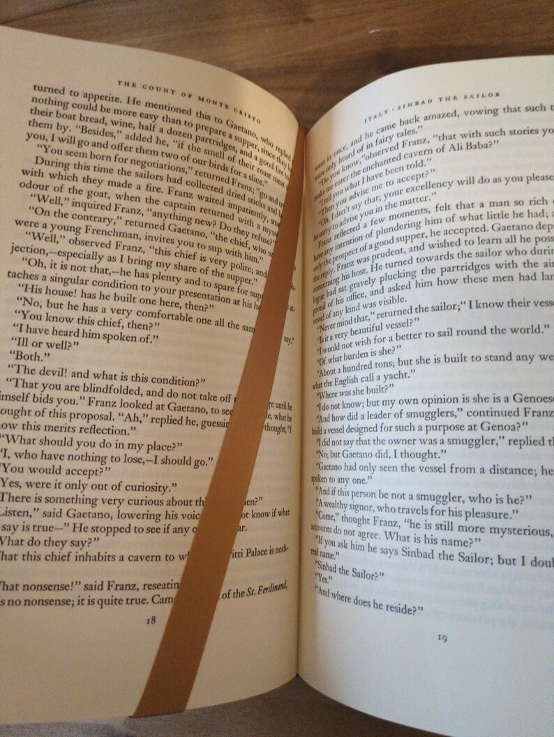 May include: Open book with text from "The Count of Monte Cristo" and "Sinbad the Sailor." A brown bookmark diagonally crosses the pages. The pages are yellowed with age.