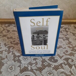 May include: A blue hardcover book titled "Self and Soul: A Woman's Guide to Enhancing Self-Esteem through Spirituality" by Adele Wilcox. The cover features a black and white photo of a woman standing on a pier overlooking a body of water.