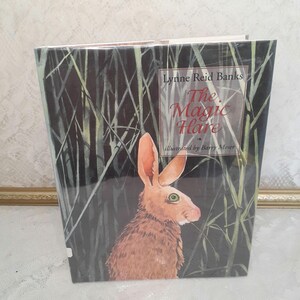 May include: The Magic Hare book cover features a brown rabbit with green eyes against a dark green background with tall grass. The title "The Magic Hare" is in white text with a black outline. The author, Lynne Reid Banks, and illustrator, Barry Moser, are also listed.