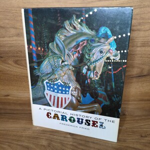 May include: A hardcover book titled A Pictorial History of the Carousel by Frederick Fried. The cover features a detailed illustration of a carousel horse adorned with a shield displaying an eagle and the American flag, set against a dark background.