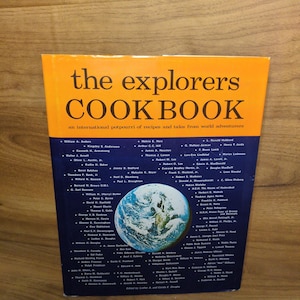 May include: A hardcover book titled The Explorers Cookbook with an orange top section and a dark blue bottom section. The cover features black text, a subtitle about international recipes, and a central circular image of the Earth from space.