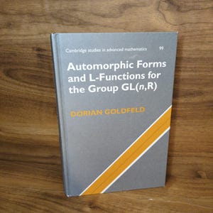 May include: A hardcover book titled "Automorphic Forms and L-Functions for the Group GL(n,R)" by Dorian Goldfeld. The book has a gray cover with white and orange diagonal stripes. The book is part of the Cambridge studies in advanced mathematics series.