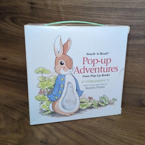 May include: A white box set of "Pop-up Adventures" books. The cover features a cartoon illustration of Peter Rabbit in a blue jacket. The text reads "Touch 'n Read" and "Four Pop-Up Books" based on Beatrix Potter's tales.