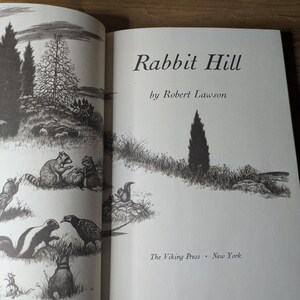 May include: The title page of the book Rabbit Hill by Robert Lawson, published by The Viking Press in New York. The page features a black and white ink illustration of a rural landscape with various animals including a raccoon, skunk, and rabbit.