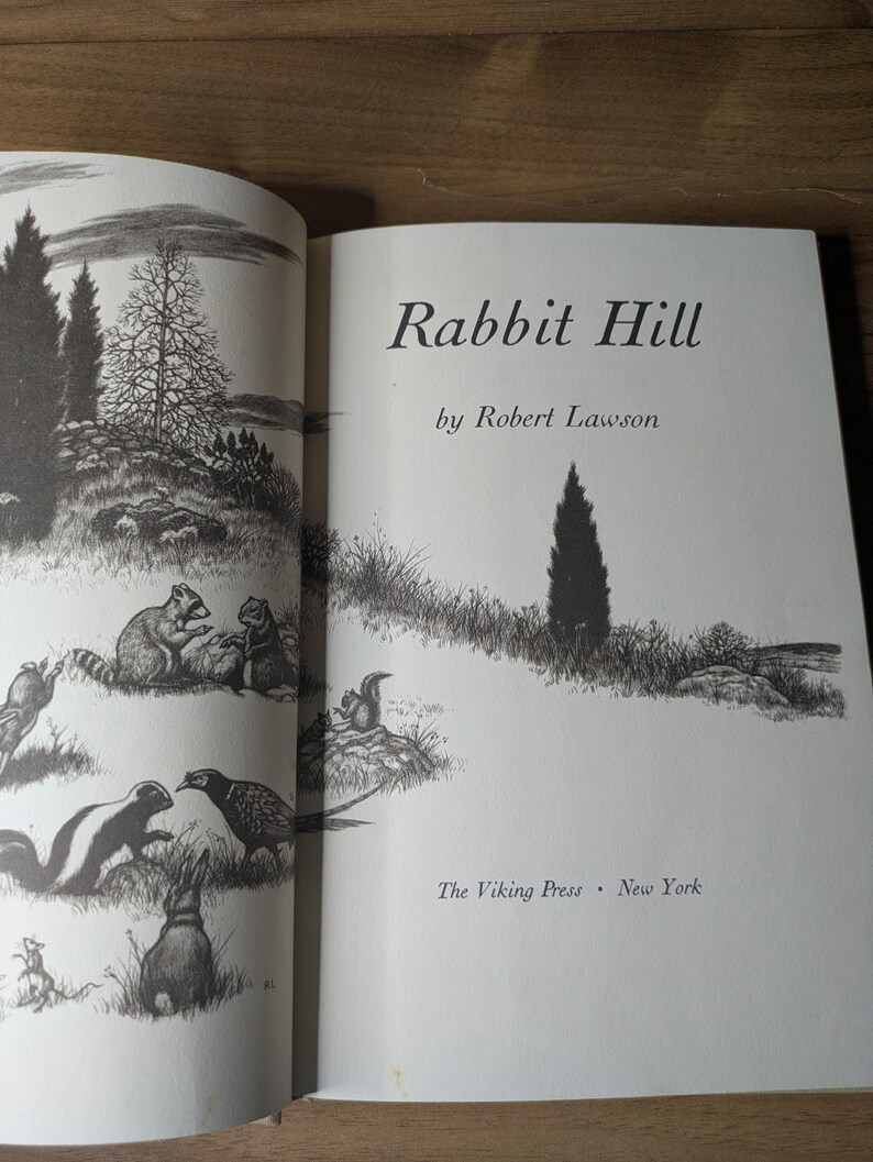 May include: The title page of the book Rabbit Hill by Robert Lawson, published by The Viking Press in New York. The page features a black and white ink illustration of a rural landscape with various animals including a raccoon, skunk, and rabbit.