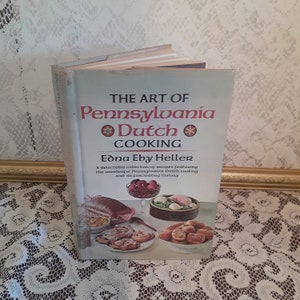 May include: A vintage cookbook titled "The Art of Pennsylvania Dutch Cooking" by Edna Eby Heller. The cover features a colorful illustration of traditional Pennsylvania Dutch food.