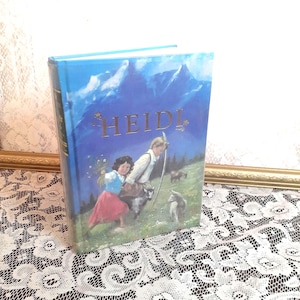 May include: A blue hardcover book with the title "HEIDI" in gold lettering. The cover features an illustration of two children, a girl and a boy, with goats in a mountain setting.