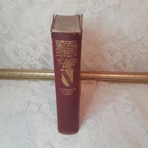 May include: A vintage burgundy book titled "Shakespeare Complete" with gold lettering and a crest design. The book's spine shows wear, suggesting its age. The book is standing upright, showcasing its title and author.