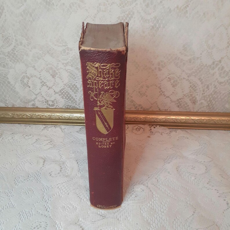 May include: A vintage burgundy book titled "Shakespeare Complete" with gold lettering and a crest design. The book's spine shows wear, suggesting its age. The book is standing upright, showcasing its title and author.