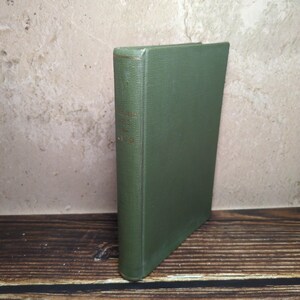 May include: A hardcover book with a solid olive green cloth cover. The spine features faint, partially obscured gold lettering. The book is standing upright, showcasing its textured fabric binding and rectangular shape in a simple, clean presentation.