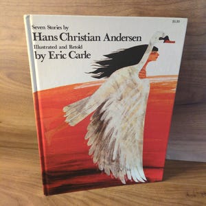 May include: A hardcover book titled "Seven Stories by Hans Christian Andersen" illustrated by Eric Carle. The cover features a swan-like figure with a red background and white text. The book is on a wooden surface.