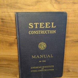 May include: A navy blue book titled "STEEL CONSTRUCTION MANUAL" with gold lettering. The cover features the AISC logo and the text "AMERICAN INSTITUTE OF STEEL CONSTRUCTION". The book is on a wooden surface.