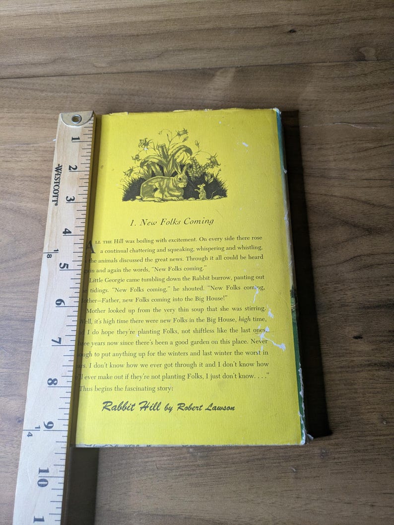 May include: A yellow book jacket for Rabbit Hill by Robert Lawson, featuring a black ink illustration of a rabbit and mouse. A wooden Westcott ruler is placed along the left edge, showing the book height is approximately 9.5 inches tall.