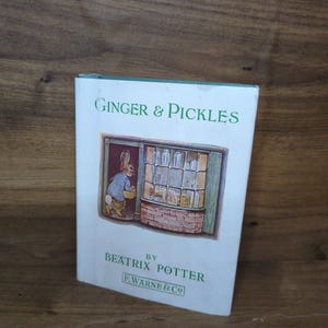 May include: A white hardcover book titled Ginger & Pickles by Beatrix Potter. The cover features a color illustration of a rabbit standing in a shop doorway. The text F. Warne & Co is printed in a rectangular box at the bottom of the book cover.