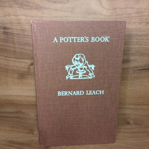 May include: A brown book titled "A Potter's Book" by Bernard Leach. The book cover features the title in light blue text, along with an illustration and the author's name. The book is standing upright.
