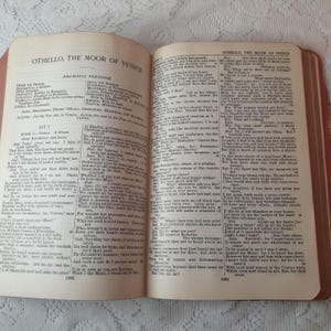 May include: An open book with aged, yellowed pages, displaying text from "Othello, the Moor of Venice." The book's cover is a faded red, and the pages are held open, revealing the text. The book is likely an antique.