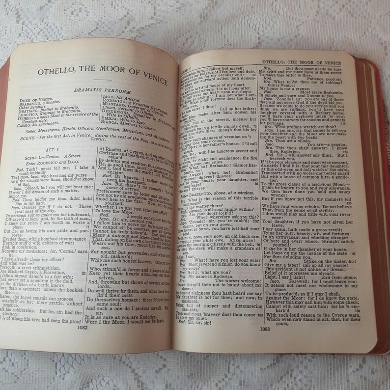 May include: An open book with aged, yellowed pages, displaying text from "Othello, the Moor of Venice." The book's cover is a faded red, and the pages are held open, revealing the text. The book is likely an antique.