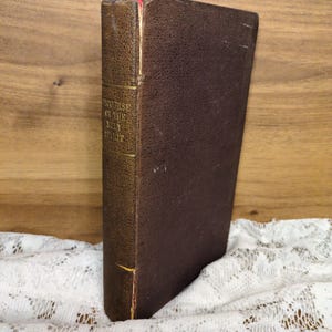 May include: A vintage hardcover book with a dark brown cover and spine. The spine features gold lettering and the title "Discourse on the Holy Spirit." A red ribbon bookmark is visible at the top. The book is placed on a white lace cloth.