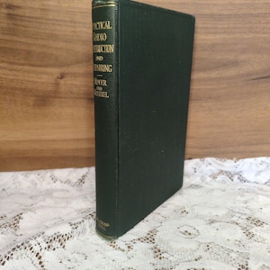 May include: A dark green hardcover book titled "Practical Radio Construction and Repairing" by Moyer and Nostrel. The book is standing upright on a white lace surface, with the spine facing the viewer. The book's title is printed in gold lettering.