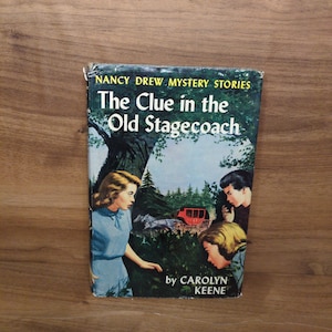 May include: Vintage hardcover book titled "The Clue in the Old Stagecoach" by Carolyn Keene. The cover features a colorful illustration of a mystery scene with three characters and a red stagecoach in a forest setting.