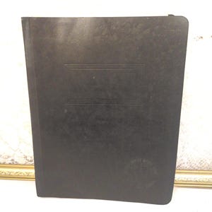 May include: A black, hardcover book with a rectangular embossed design on the front cover. The book has a rounded spine and a small, circular emblem near the bottom right corner. The book is standing upright.