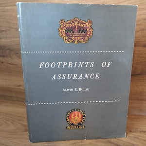 May include: A hardcover book titled "Footprints of Assurance" by Alwin E. Bulau. The book has a gray cover with decorative emblems and text in white. The top emblem reads "Insurance" and the bottom emblem reads "Department & Tamar Fire Insurance".