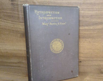 May include: A dark gray hardcover book titled Retrospection and Introspection by Mary Baker G. Eddy. The front cover features gold embossed lettering and a circular gold emblem in the center with text and a sunburst design. The book has a rectangular shape.