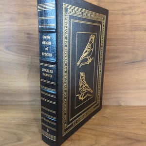 May include: A hardcover book titled "On the Origin of Species" by Charles Darwin. The book has a black cover with gold lettering and illustrations of birds. The spine also features gold lettering. The book is displayed upright.
