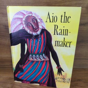 May include: A hardcover book titled Aio the Rain-maker by Fiona French. The cover features a central illustration of a figure wearing a dark brown mask, a large pink headdress, and a dress with vertical red and blue stripes, accented by a belt of skulls.