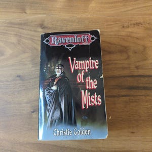 Puede incluir: Un libro de bolsillo titulado "Vampire of the Mists" de Christie Golden. La portada presenta una imagen oscura de un vampiro con una capa negra. El libro es parte de la serie Ravenloft Books.