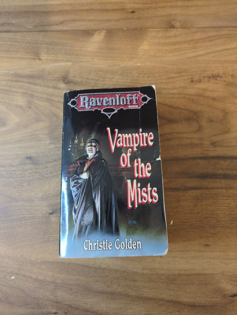 Puede incluir: Un libro de bolsillo titulado "Vampire of the Mists" de Christie Golden. La portada presenta una imagen oscura de un vampiro con una capa negra. El libro es parte de la serie Ravenloft Books.