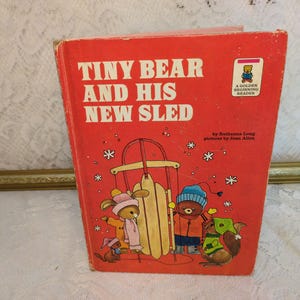 May include: A vintage children's book titled "Tiny Bear and His New Sled" with a red cover. The cover features illustrations of cartoon animals and a sled. The book is part of the "Golden Beginning Reader" series.