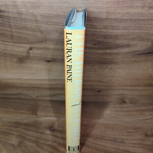 May include: A book with a yellow spine. The title "THE HIERARCHY OF HELL" and the author's name, LAURAN PAINE, are printed in black. The top edge is light blue. The book is set against a wooden background.