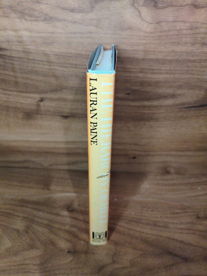 May include: A book with a yellow spine. The title "THE HIERARCHY OF HELL" and the author's name, LAURAN PAINE, are printed in black. The top edge is light blue. The book is set against a wooden background.