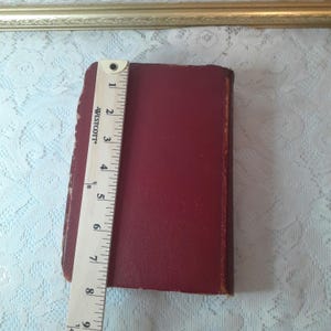 May include: A burgundy-colored book with a leather-like cover is measured with a wooden ruler. The ruler shows the book is approximately 8 inches long. The book is on a white lace surface.