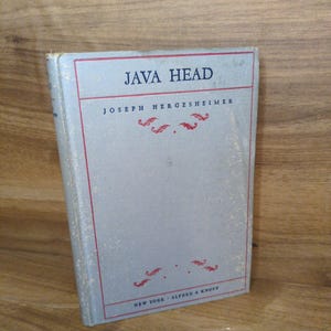 May include: A hardcover book titled Java Head by Joseph Hergesheimer, published by Alfred A. Knopf in New York. The book features a light gray cover with a thin red rectangular border, red decorative flourishes, and dark blue printed text.