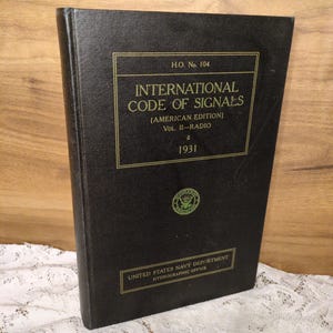 May include: A black book titled "INTERNATIONAL CODE OF SIGNALS" (American Edition), Vol. II-Radio, 1931. The cover features gold lettering and a seal from the United States Navy Department Hydrographic Office. The book is standing upright.