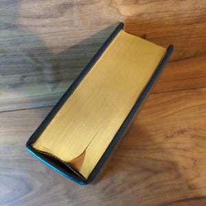 May include: A hardcover book with gold-gilded edges and a dark blue cover. The book is lying on a wooden surface, showcasing its rectangular shape and the contrast between the gold and the dark cover.