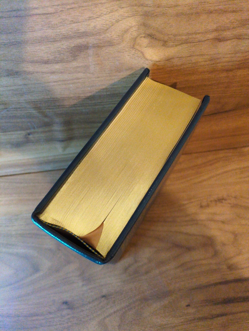 May include: A hardcover book with gold-gilded edges and a dark blue cover. The book is lying on a wooden surface, showcasing its rectangular shape and the contrast between the gold and the dark cover.
