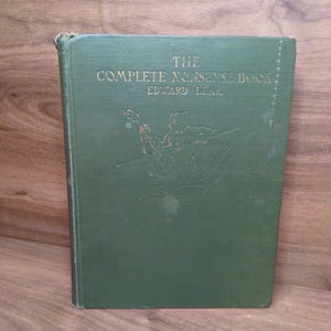 May include: A green hardcover book titled The Complete Nonsense Book by Edward Lear. The front cover features gold embossed lettering and a central illustration of a cat playing a stringed instrument while seated in a small boat on water.