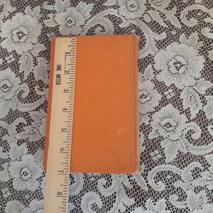K&ouml;nnte beinhalten: Ein orangefarbenes Buch mit einem Lineal daneben. Das Lineal zeigt, dass das Buch etwa 20 cm lang ist. Das Buch liegt auf einer wei&szlig;en Spitzentischdecke.