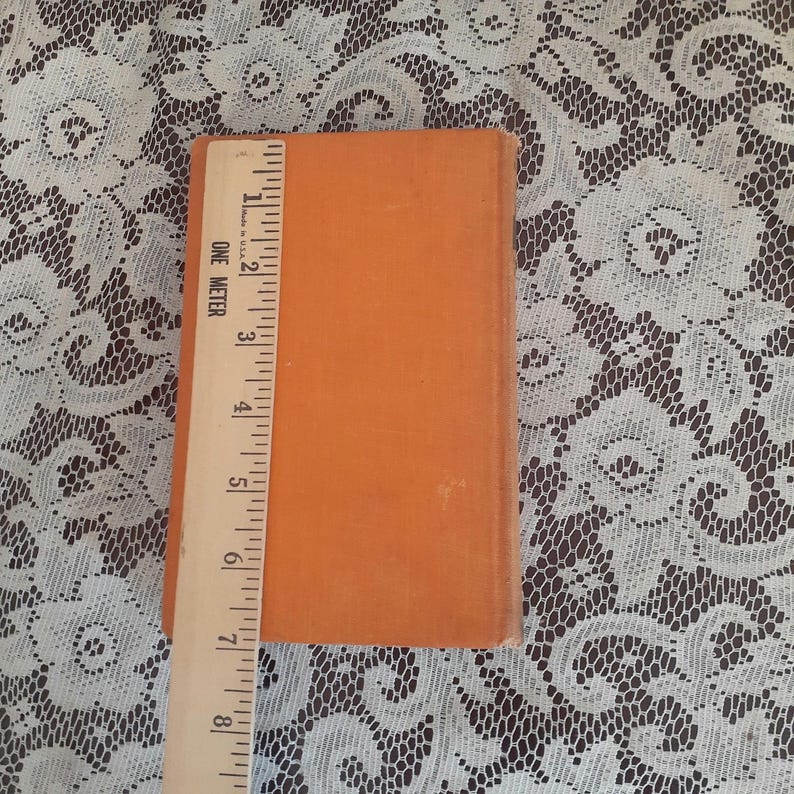 K&ouml;nnte beinhalten: Ein orangefarbenes Buch mit einem Lineal daneben. Das Lineal zeigt, dass das Buch etwa 20 cm lang ist. Das Buch liegt auf einer wei&szlig;en Spitzentischdecke.