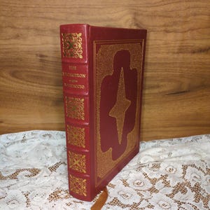 May include: A hardcover book titled "The Decameron" by Boccaccio. The book has a red cover with gold embossed designs and text. A gold starburst design is on the front cover. A brown leather bookmark is visible.