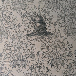 May include: A beige fabric featuring a repeating black line art pattern of a rabbit sitting upright among clusters of bell-shaped flowers and leafy stems. The rabbit is positioned centrally, surrounded by detailed botanical illustrations.