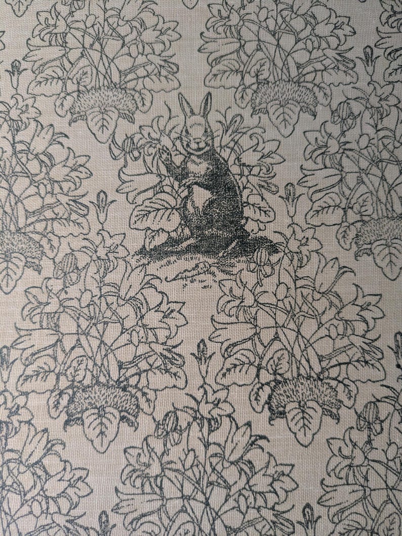 May include: A beige fabric featuring a repeating black line art pattern of a rabbit sitting upright among clusters of bell-shaped flowers and leafy stems. The rabbit is positioned centrally, surrounded by detailed botanical illustrations.