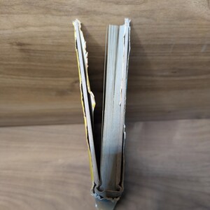 May include: A book with a yellow cover and a damaged, detached spine showing the internal binding and paper pages. The cover edges are frayed and worn, revealing the structural layers of the book's construction from a top-down perspective.