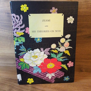May include: A book titled "ZEAMI and HIS THEORIES ON NOH" with a black cover. The cover features colorful floral illustrations, including white, red, and yellow flowers. The book is standing upright on a wooden surface.
