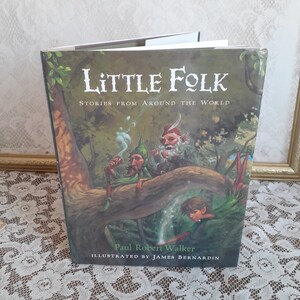 May include: A book titled "Little Folk: Stories from Around the World" by Paul Robert Walker, illustrated by James Bernardin. The cover features a whimsical illustration of three fairies in a forest setting.