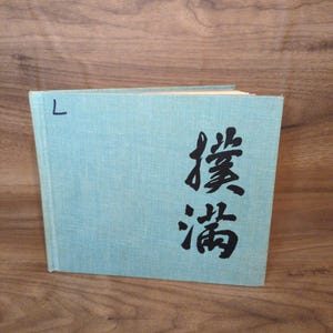 May include: A light blue hardcover book with a textured fabric cover. The front cover features the black ink calligraphy characters 撲滿 and the handwritten letter L in the upper left corner. The book is positioned upright with visible pages.