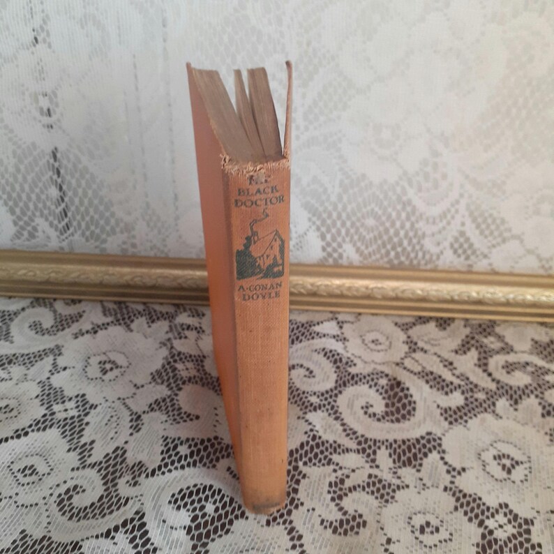 K&ouml;nnte beinhalten: Ein Vintage-Buch mit dem Titel "The Black Doctor" von A. Conan Doyle. Das Buch hat einen verblichenen orangefarbenen Einband mit schwarzem Text und einer kleinen Illustration eines Hauses. Die Seiten sind gealtert, und der Buchr&uuml;cken weist Abnutzungserscheinungen auf.